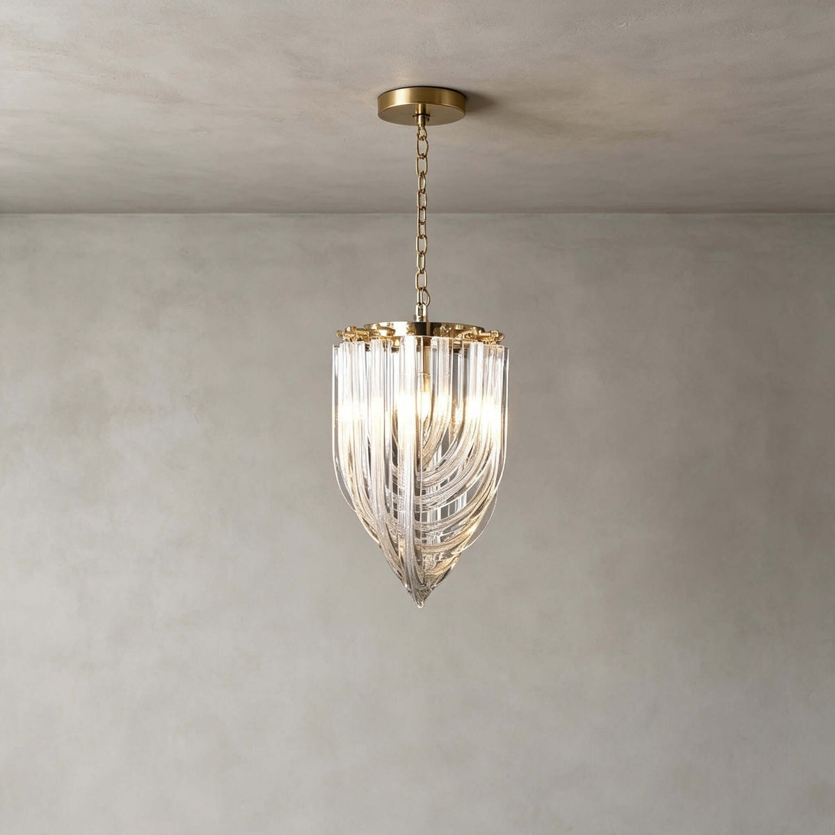 The smallest Clérachic pendant, with a slim glass-rod body ending in a point, hangs on a fine gold chain against a grey plaster wall.