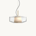 A Clérachic pendant with a wide clear glass shade, a pleated white inner shade and a brass stem, on a plain white backdrop.
