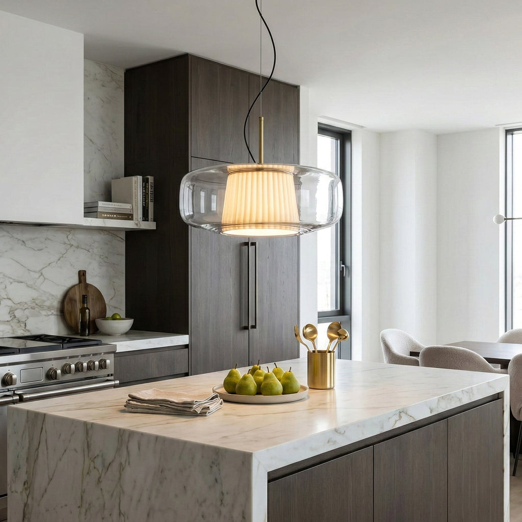 Clear glass Clérachic pendant glowing over a marble kitchen island, with dark wood cabinets and a bowl of pears below.