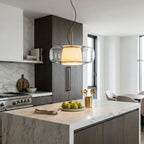 Clear glass Clérachic pendant glowing over a marble kitchen island, with dark wood cabinets and a bowl of pears below.