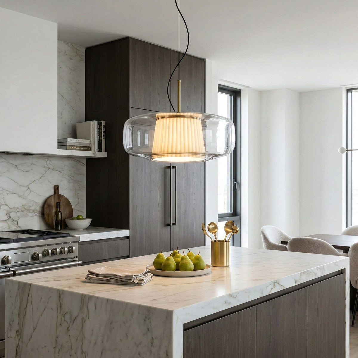 Clear glass Clérachic pendant glowing over a marble kitchen island, with dark wood cabinets and a bowl of pears below.