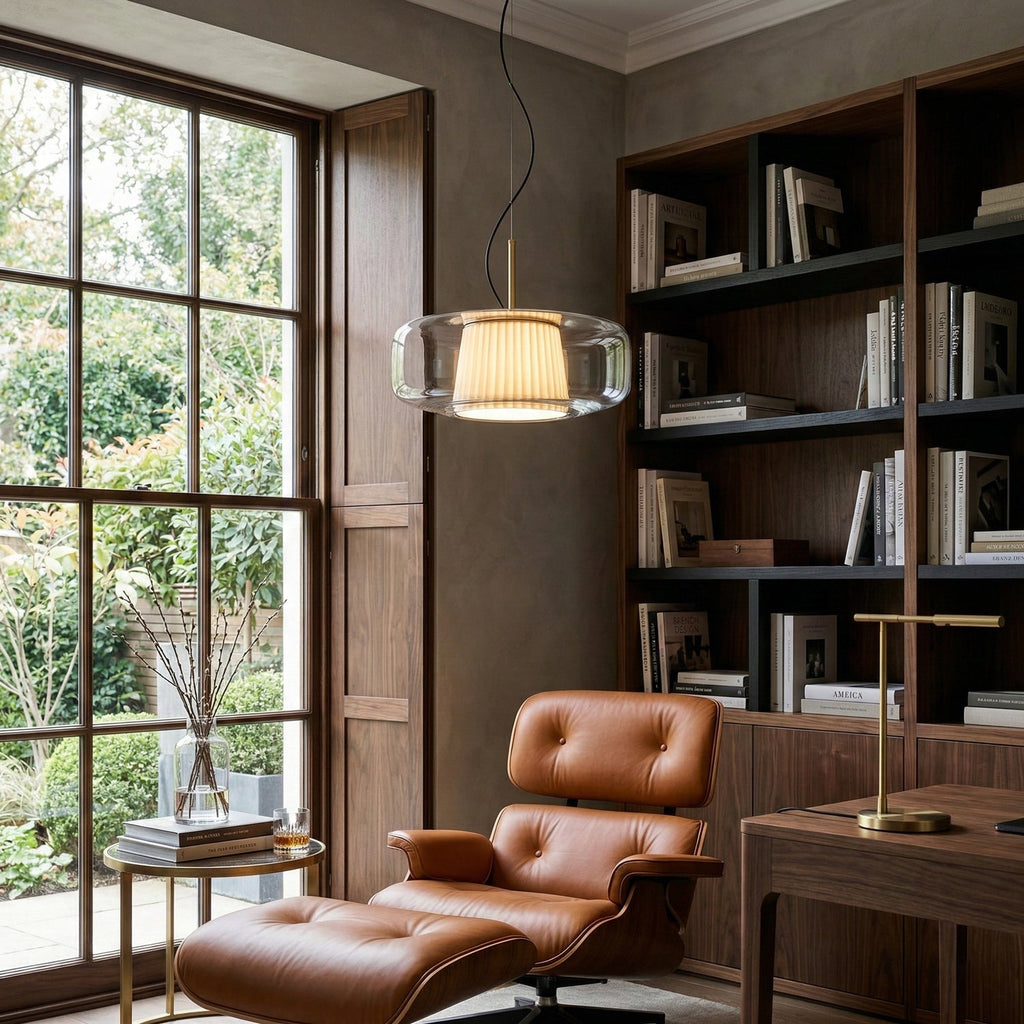 A Clérachic clear glass pendant lit beside a tall window, with dark bookshelves and a tan leather armchair in the corner of the room.