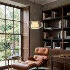 A Clérachic clear glass pendant lit beside a tall window, with dark bookshelves and a tan leather armchair in the corner of the room.
