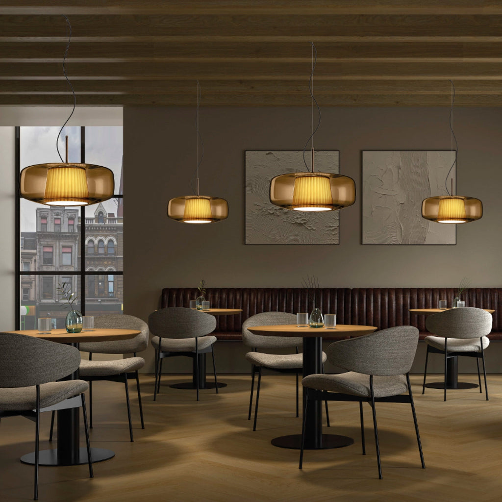 Four amber glass Clérachic pendants hang at different heights over café tables in a restaurant with grey chairs and a wooden ceiling.