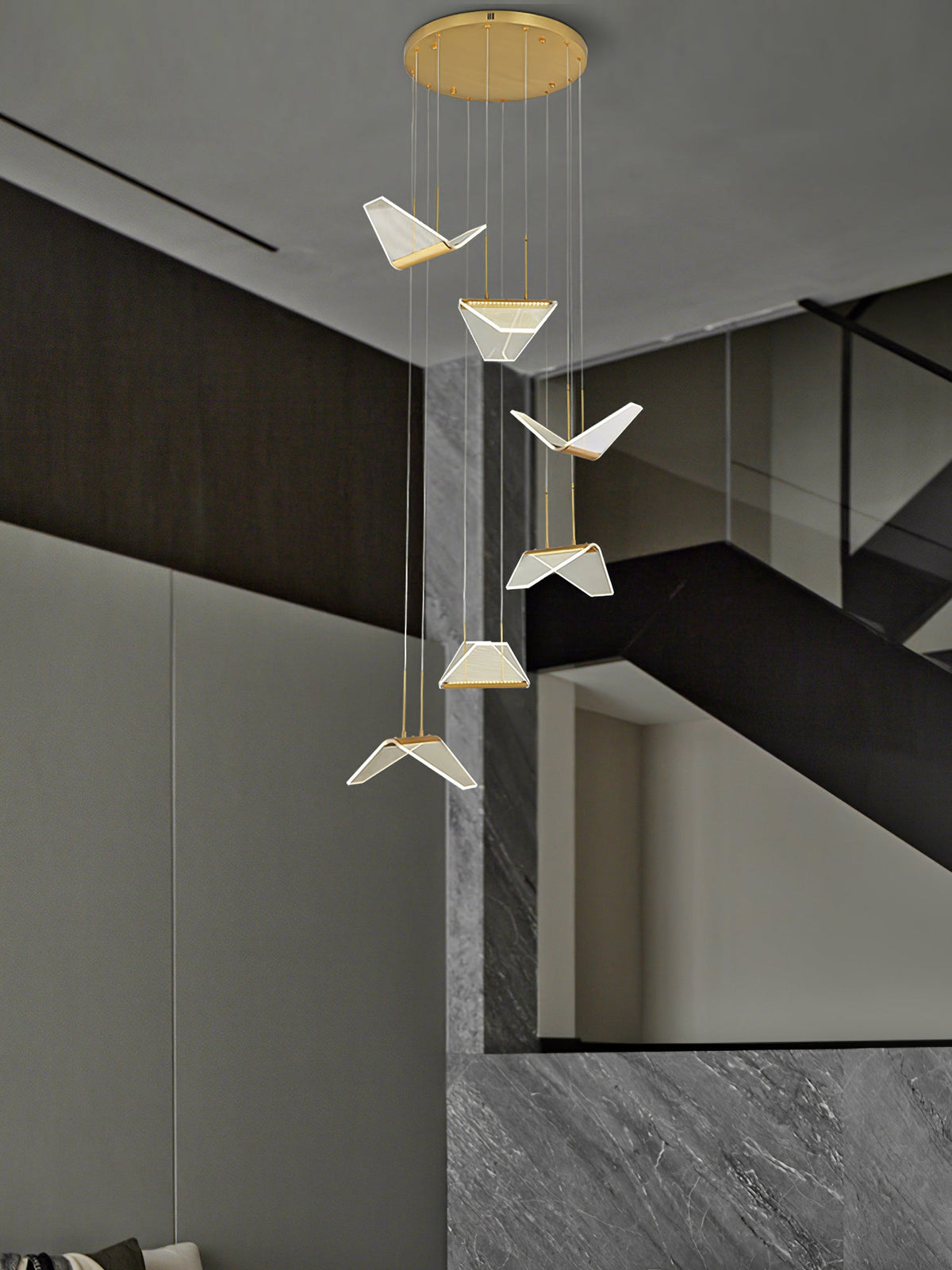 Pale folded shades of this Clérachic pendant light descend from a round gold plate beside a grey stone staircase.