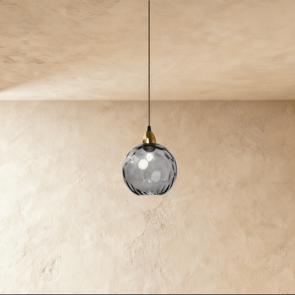 Clérachic gold pendant light with a smoky gray blown glass globe, hung from a brass fitting against a plaster wall.