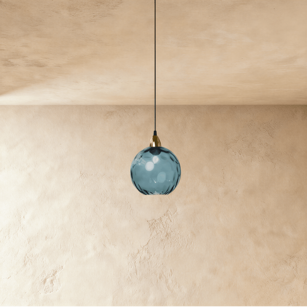 Clérachic gold pendant light in a blue blown glass globe, suspended on a slim black cord over a plaster wall.