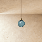 Clérachic gold pendant light in a blue blown glass globe, suspended on a slim black cord over a plaster wall.