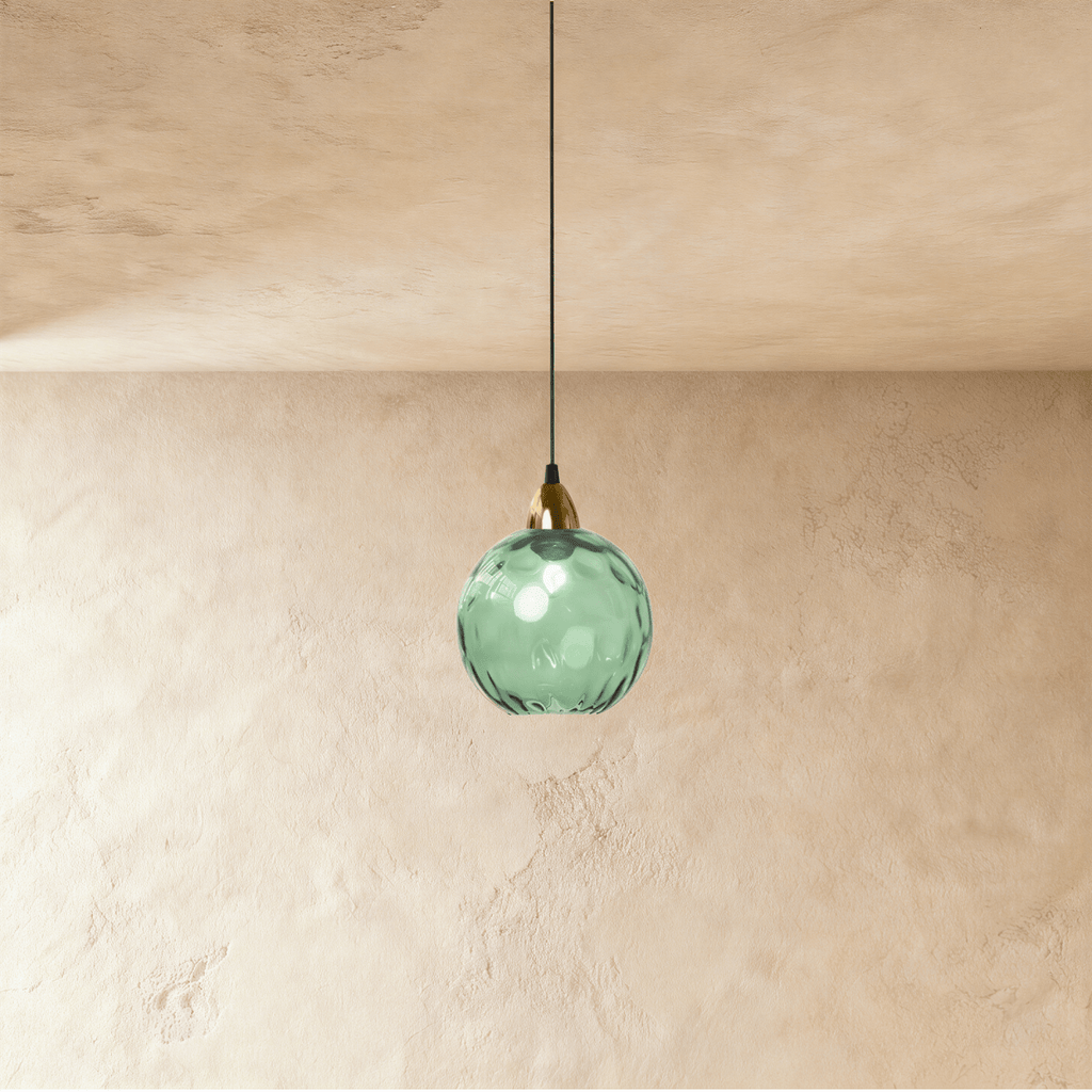 Clérachic gold pendant light with a green blown glass globe hanging from a brass cap against a plaster wall.