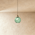 Clérachic gold pendant light with a green blown glass globe hanging from a brass cap against a plaster wall.