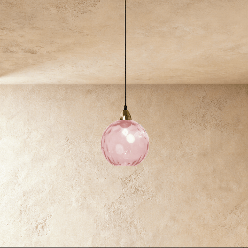 Clérachic gold pendant light with a pink blown glass globe suspended from a brass fitting on a plaster wall.