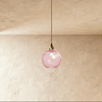 Clérachic gold pendant light with a pink blown glass globe suspended from a brass fitting on a plaster wall.