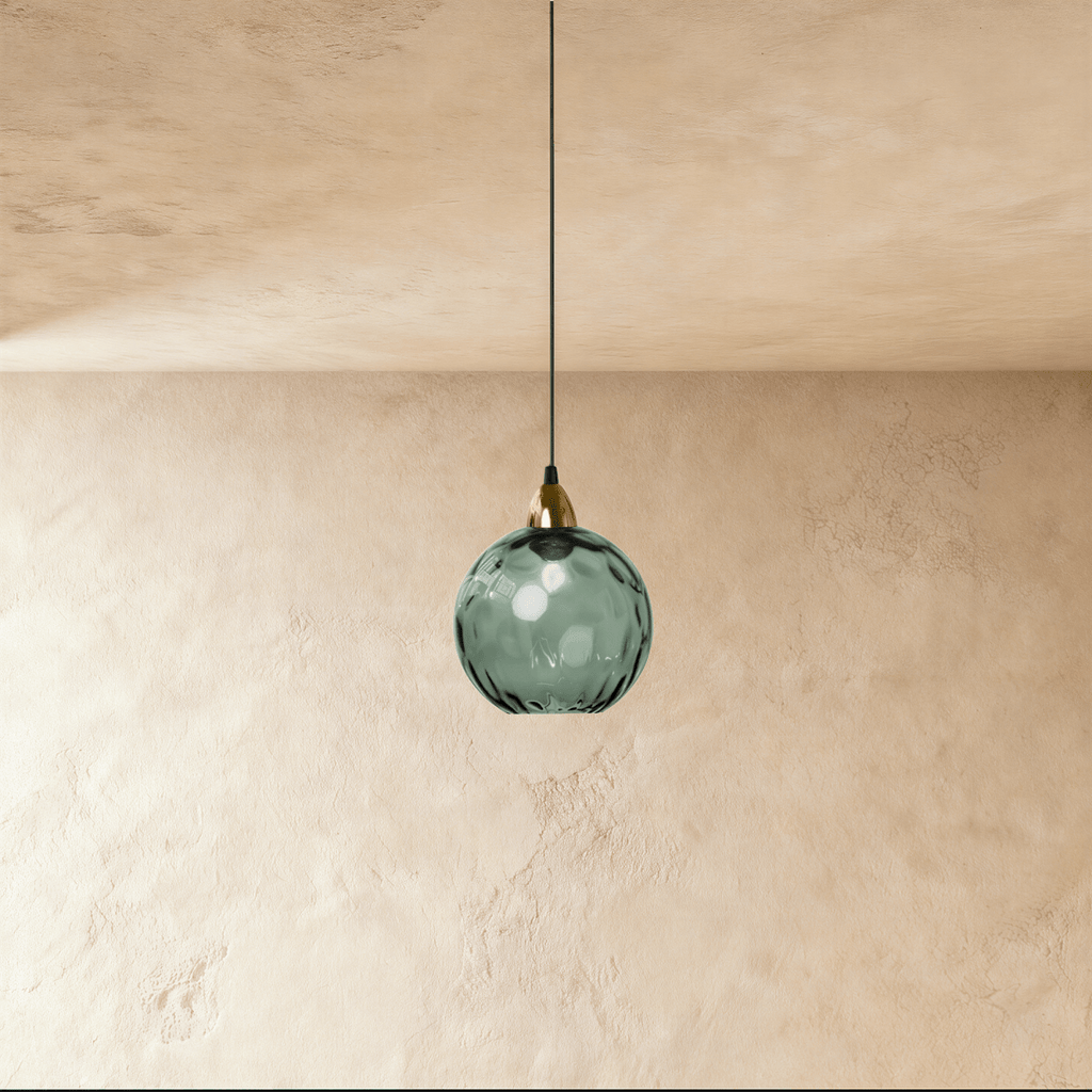 Clérachic gold pendant light with a blackish green blown glass globe hanging from a slim cord on a plaster wall.