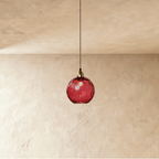 Clérachic gold pendant light with a red blown glass globe, its dimpled surface glowing above a plaster wall.