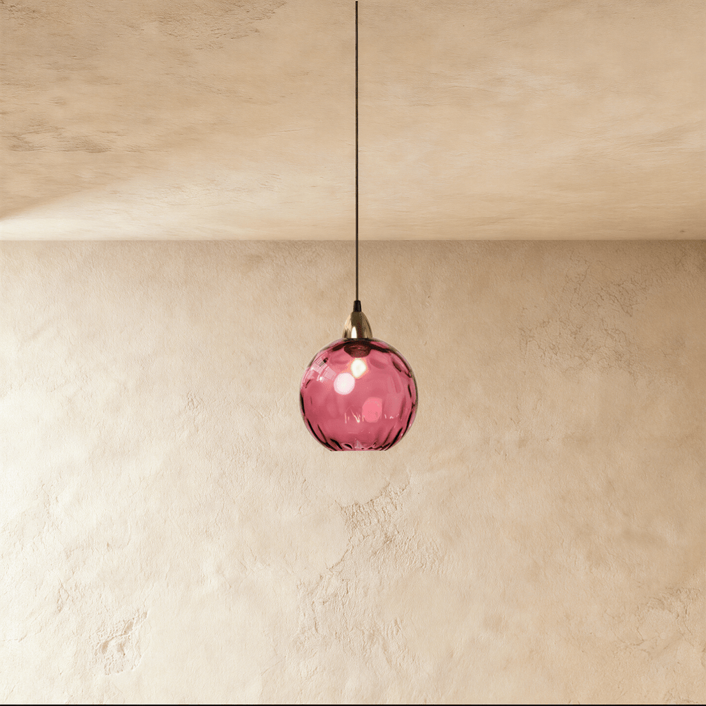 Clérachic gold pendant light with a rose red blown glass globe suspended from a brass cap on a plaster wall.