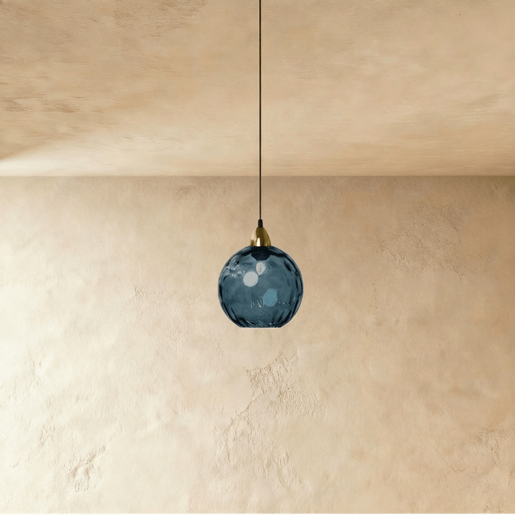 Clérachic gold pendant light with a dark blue blown glass globe hanging from a brass fitting on a plaster wall.