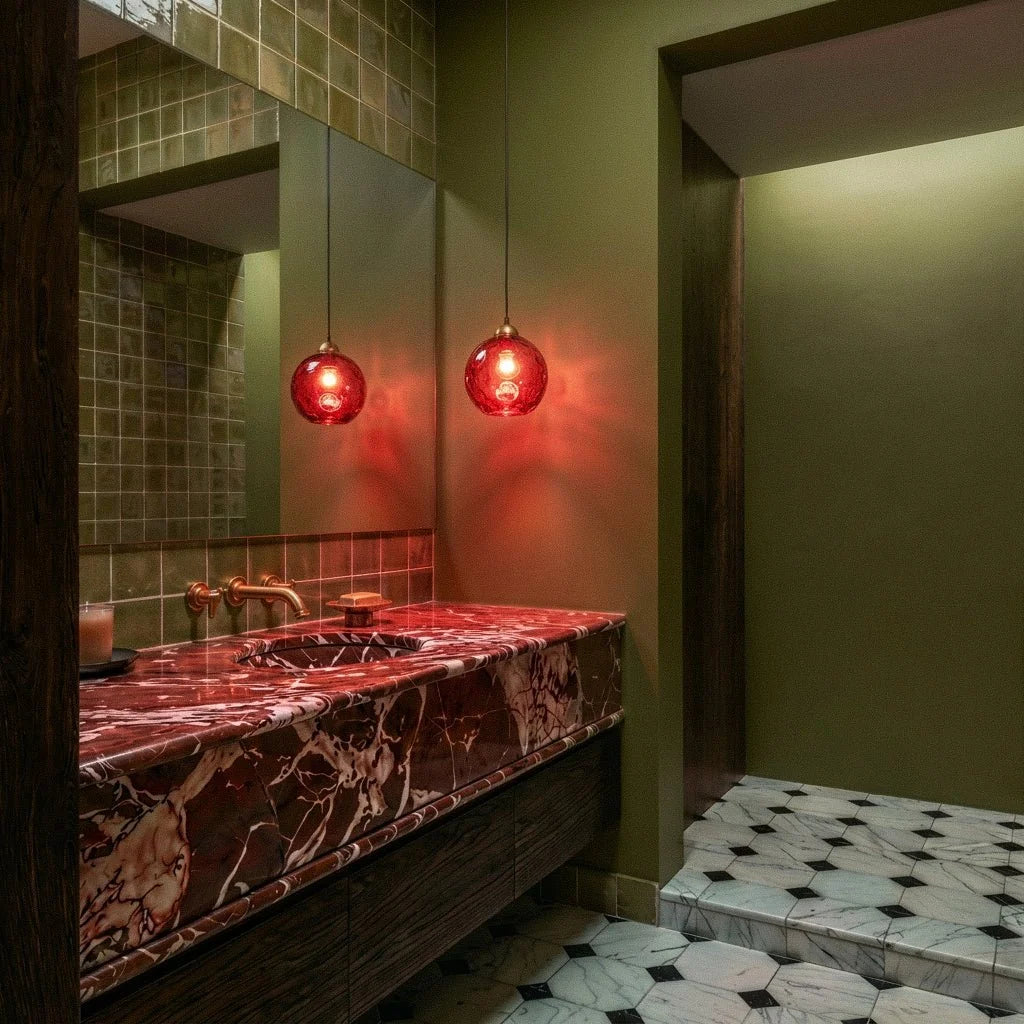 Clérachic gold pendant light with a red blown glass globe reflected in a bathroom mirror above a marble vanity.