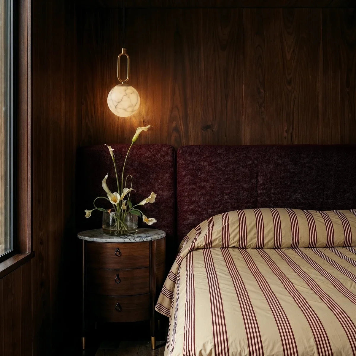 Clérachic gold alabaster pendant glowing above a bedside table, set against a dark wood-panelled bedroom wall.