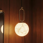 Clérachic pendant light detail, its gold oval link and glowing alabaster globe reflected in a nearby mirror.
