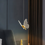 Clérachic brass butterfly pendant light glowing warm white beside a velvet armchair in a dim bedroom.
