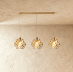Clérachic three clear glass petal pendants in a row from a long brass bar canopy, hung against limewash.