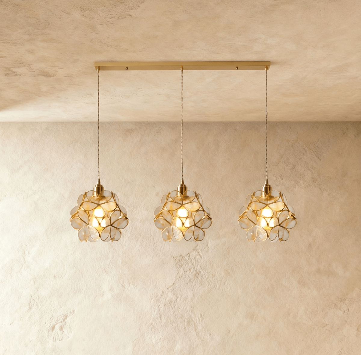 Clérachic three clear glass petal pendants in a row from a long brass bar canopy, hung against limewash.