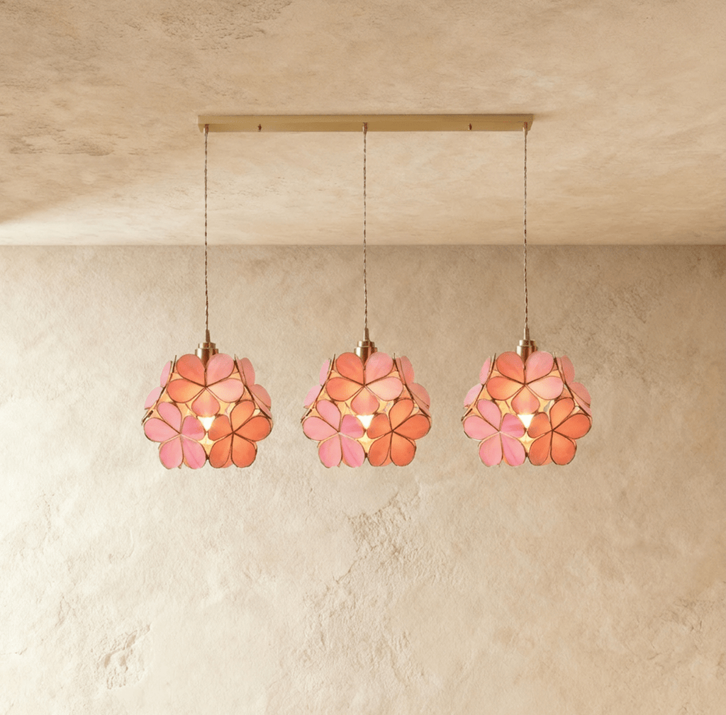Clérachic three pink glass petal pendants in a row from a long brass bar canopy, hung against limewash.