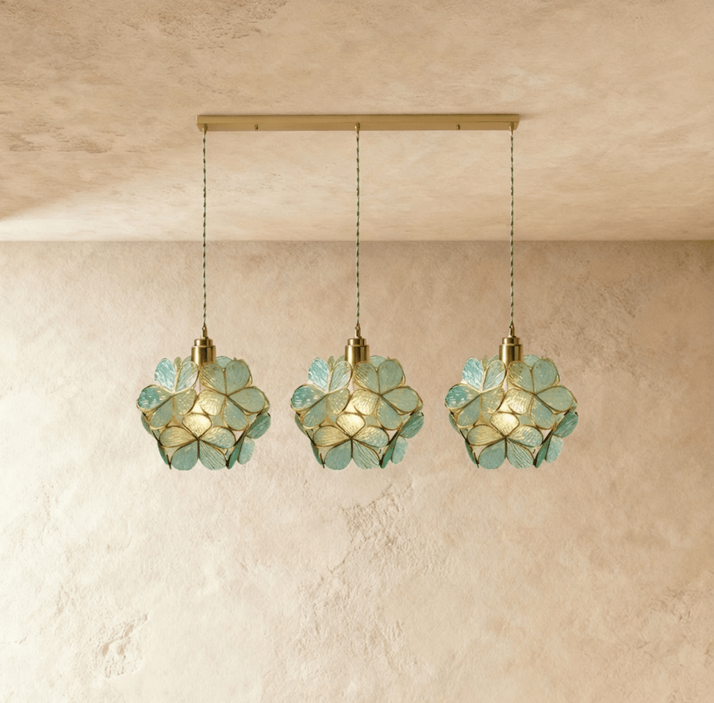Clérachic three blue-green glass petal pendants in a row from a long brass bar canopy, hung against limewash.