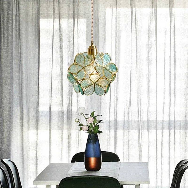 Clérachic blue-green glass petal pendant over a white dining table with a navy vase, soft sheer curtains behind.