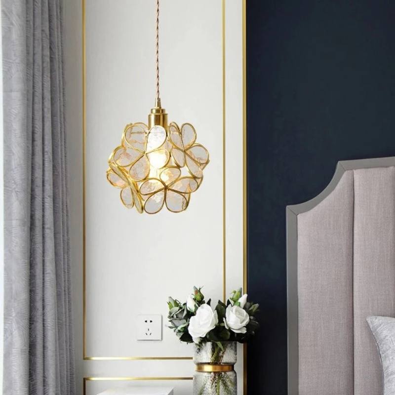Clérachic clear glass petal pendant edged in brass beside a navy panel and a grey headboard, above white roses in a vase.