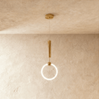 Clérachic gold pendant with a glowing opal ring held by a brass stem and small ball, hung on a fine wire against plaster.