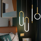Clérachic gold pendants in U, rod and ring shapes glowing beside a bed with an orange headboard and dark green wall.