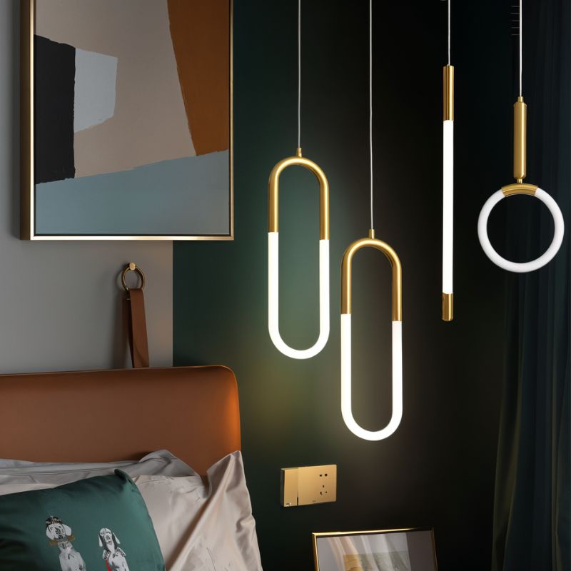 Clérachic gold pendants in U, rod and ring shapes glowing beside a bed with an orange headboard and dark green wall.