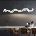 Lit Clérachic pendant with a rippling wave shape above a pale kitchen island, with brown quilted bar stools below against a dark grey wall.