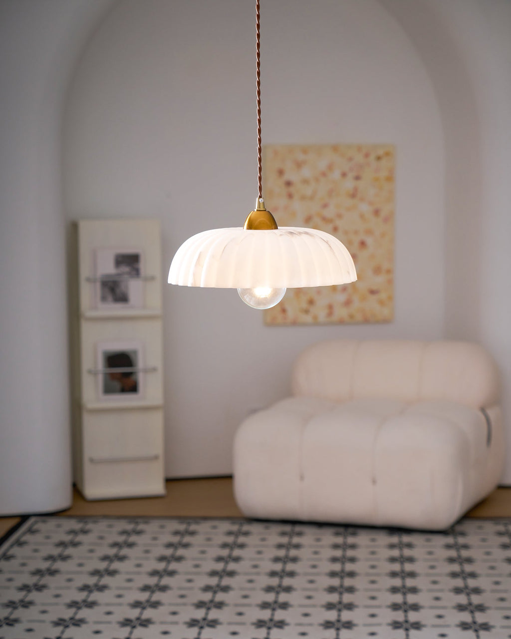 Lit ribbed white Clérachic pendant in a pale arched alcove, with a cream bouclé armchair and a peach abstract painting.