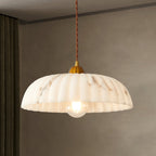 Lit Clérachic pendant with a ribbed marbled shade and a gold cap, hanging in front of a taupe wall and a dark curtain.