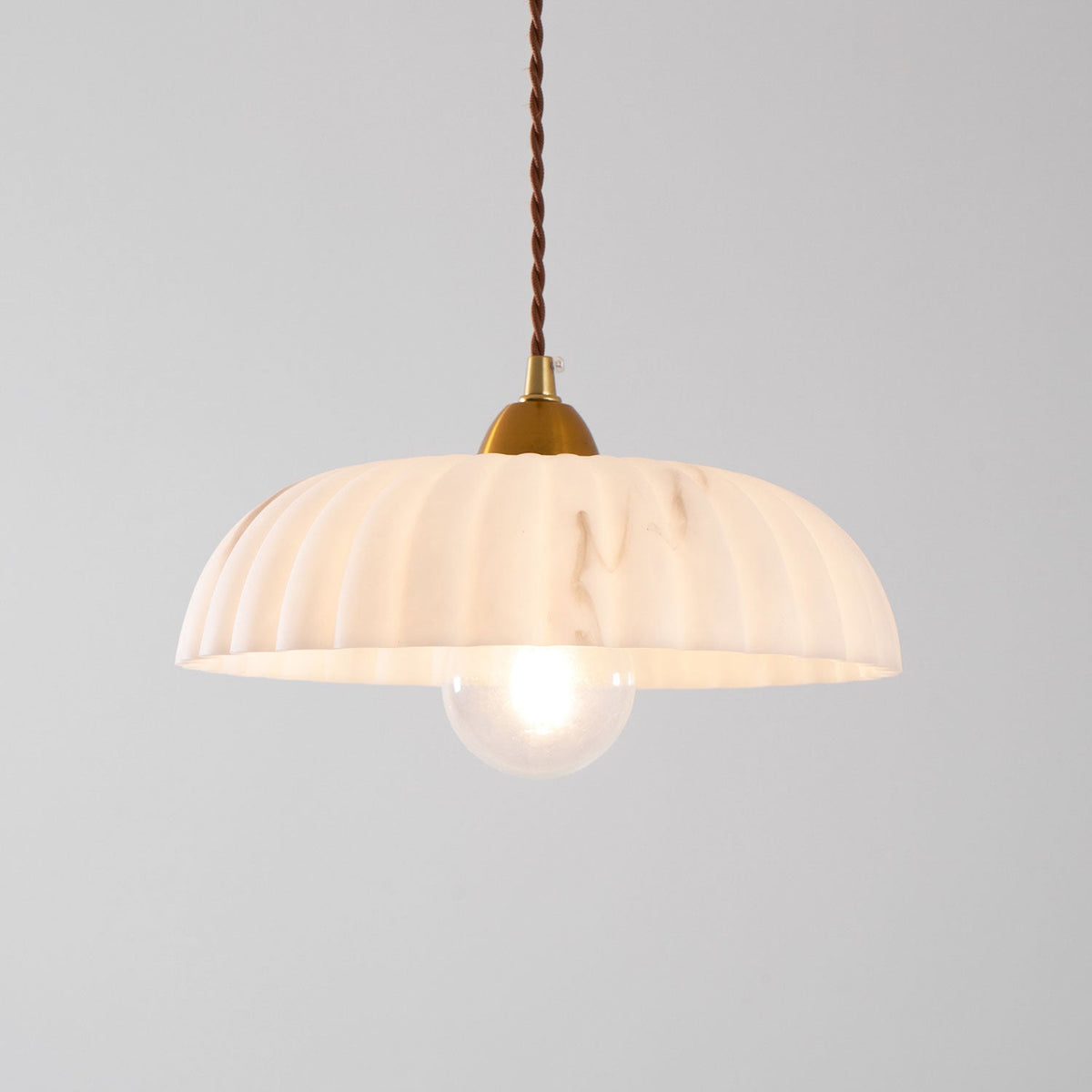 A Clérachic pendant with a peachy-white ribbed shade and a clear glass bulb, glowing on a pale grey backdrop.