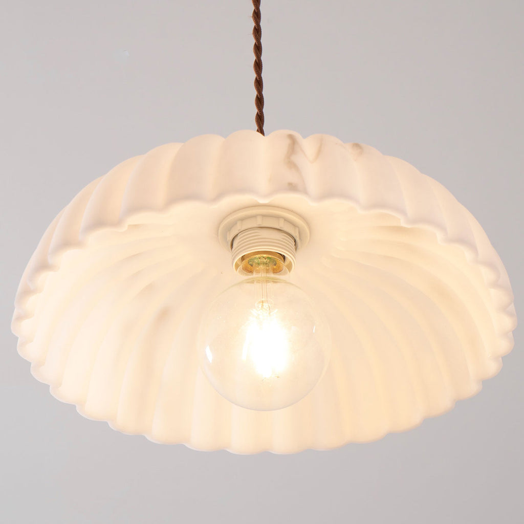 View from below of a lit, scalloped shade on this Clérachic pendant, with a ceramic socket and a round bulb glowing at the centre.