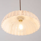 View from below of a lit, scalloped shade on this Clérachic pendant, with a ceramic socket and a round bulb glowing at the centre.