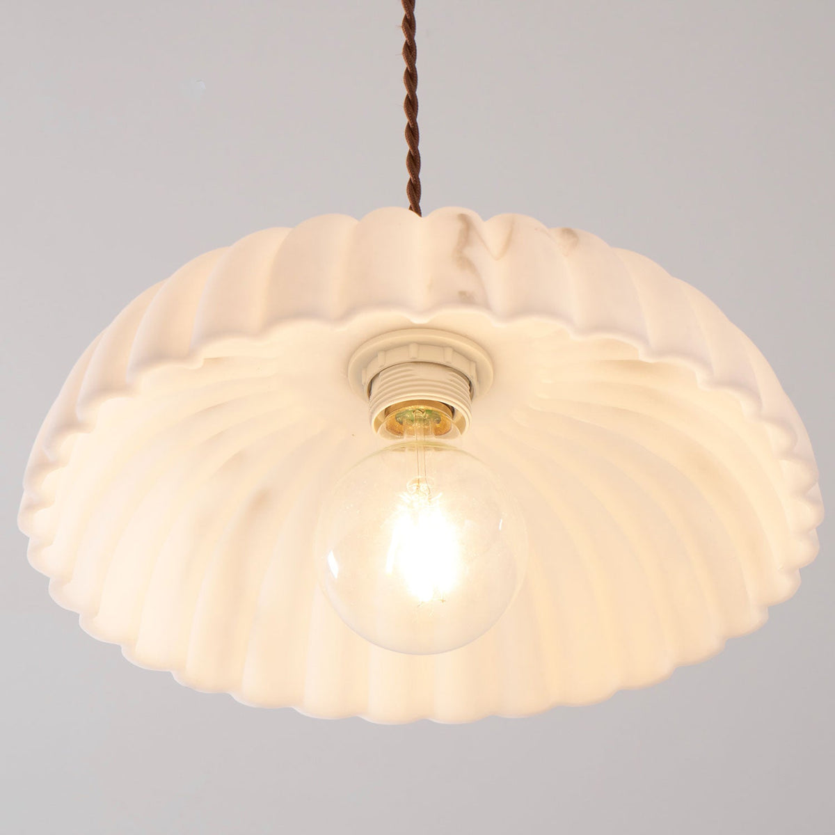 View from below of a lit, scalloped shade on this Clérachic pendant, with a ceramic socket and a round bulb glowing at the centre.
