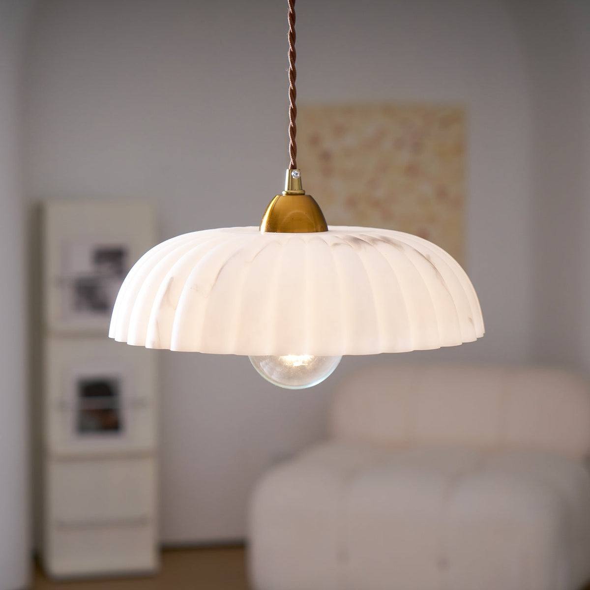 A Clérachic pendant with a ribbed white shade and a clear bulb, hanging in front of a cream chair and a peach painting.