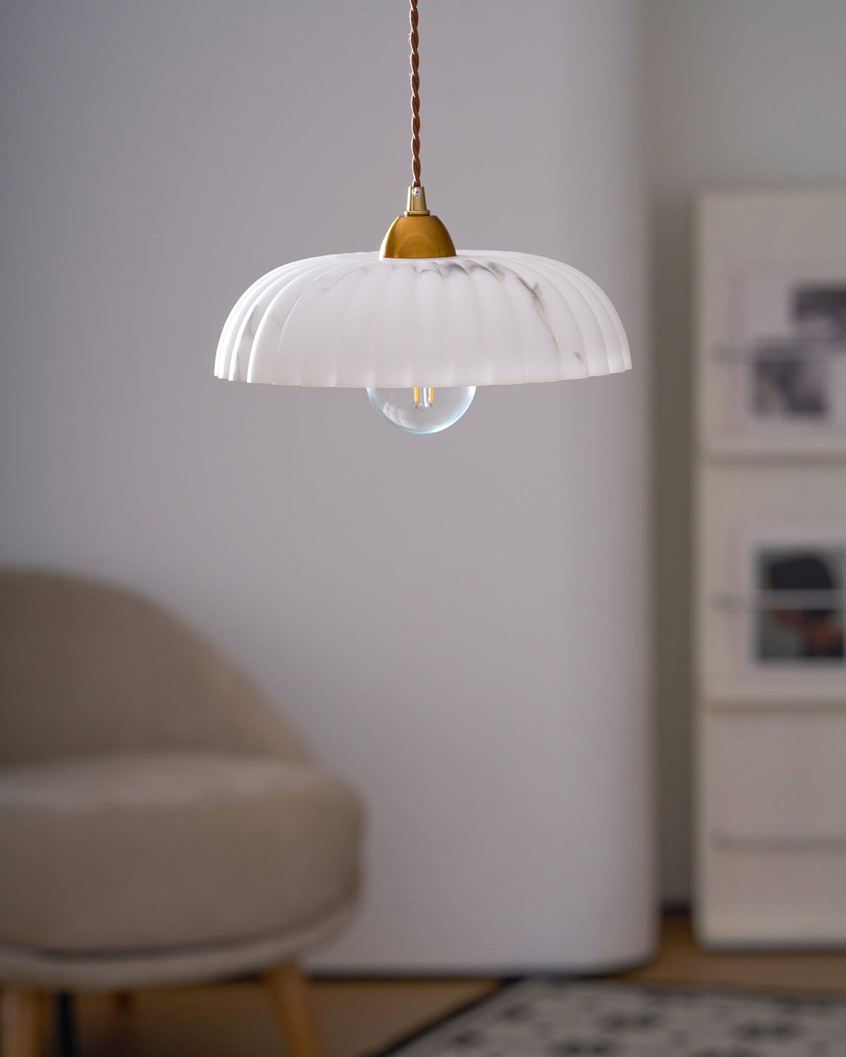 A Clérachic pendant with a white ribbed shade and a visible bulb, in front of a rounded wall and a tan armchair.