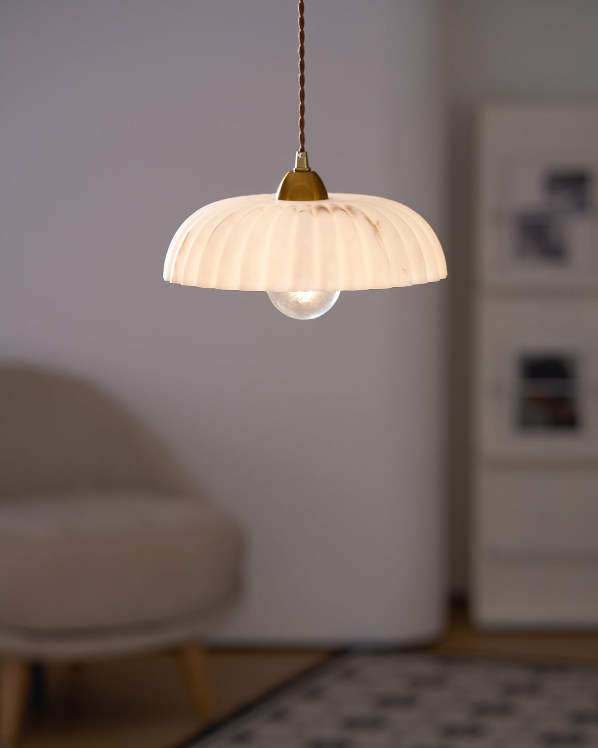 Lit Clérachic pendant with a warm glowing ribbed shade, in front of a soft-focus tan armchair and a pale wall.