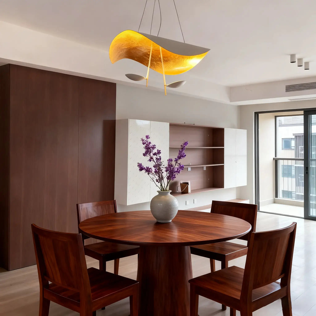A gold-and-grey Clérachic pendant light hangs over a round wood dining table set with a vase of purple flowers.