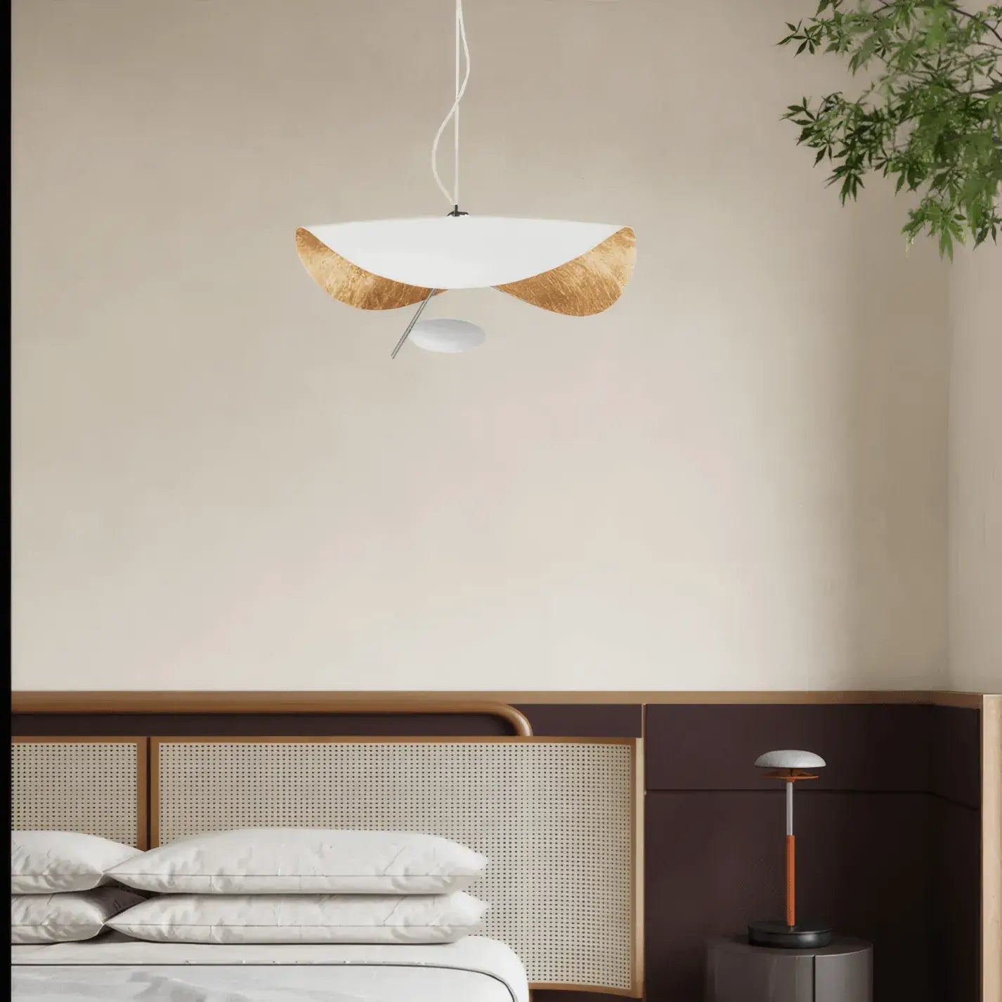 The Clérachic pendant light in warm white and gold, hung above a bed with a woven cane headboard and bedside plant.