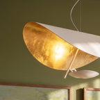 Close view of the Clérachic white pendant: sunlit gold-leaf underside of the shade, a slim brass arm and a pale reflecting disc