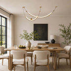 Two lit curved strips of this Clérachic pendant above a wooden table, with a dark framed painting and a tall window to the left.