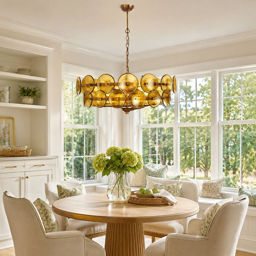 Green and gold disc chandelier from Clérachic hanging above a round pedestal dining table in a bright breakfast nook with tall windows.