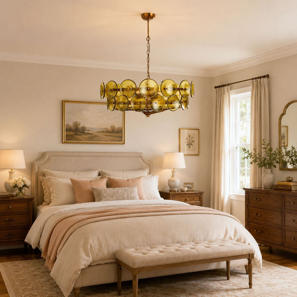 This Clérachic chandelier in green and gold glass discs hangs at the foot of a bedroom, above a cream bed and a tufted bench.