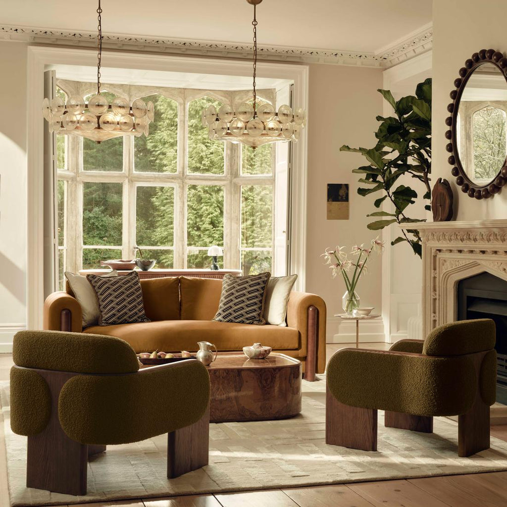 Two white glass disc chandeliers hang side by side in front of a large bay window, over a camel sofa and two olive boucle chairs; Clérachic.